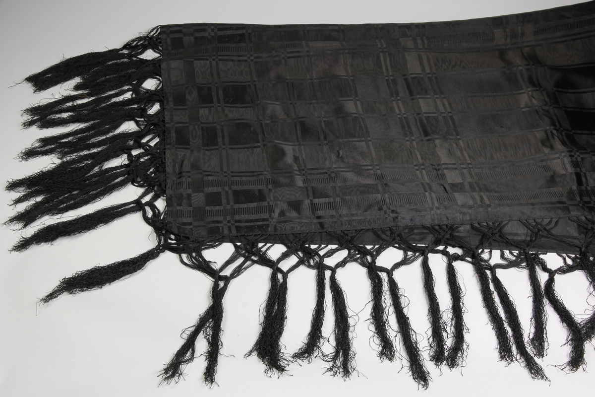 Svart sidensjal med knytna krusade fransar. Sjalens mönster består av rutor i olika storlekar och vävtekniker, blanka rutor i satin och matt i rips som är moirebehandlade.