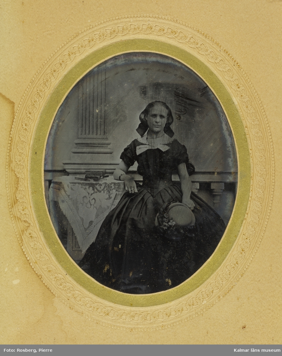 Porträtt av fröken Josefine Strandberg, nästan helbild, hon sitter vid ett bord, i sitt knä har hon en hatt med blommor på.