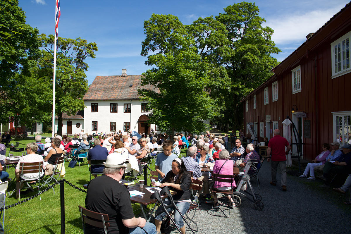 Uteområde på Gjøvik gård, med den hvite hovedbygningen og den røde Drengestua. Mange mennesker rundt kafebord. Sol.