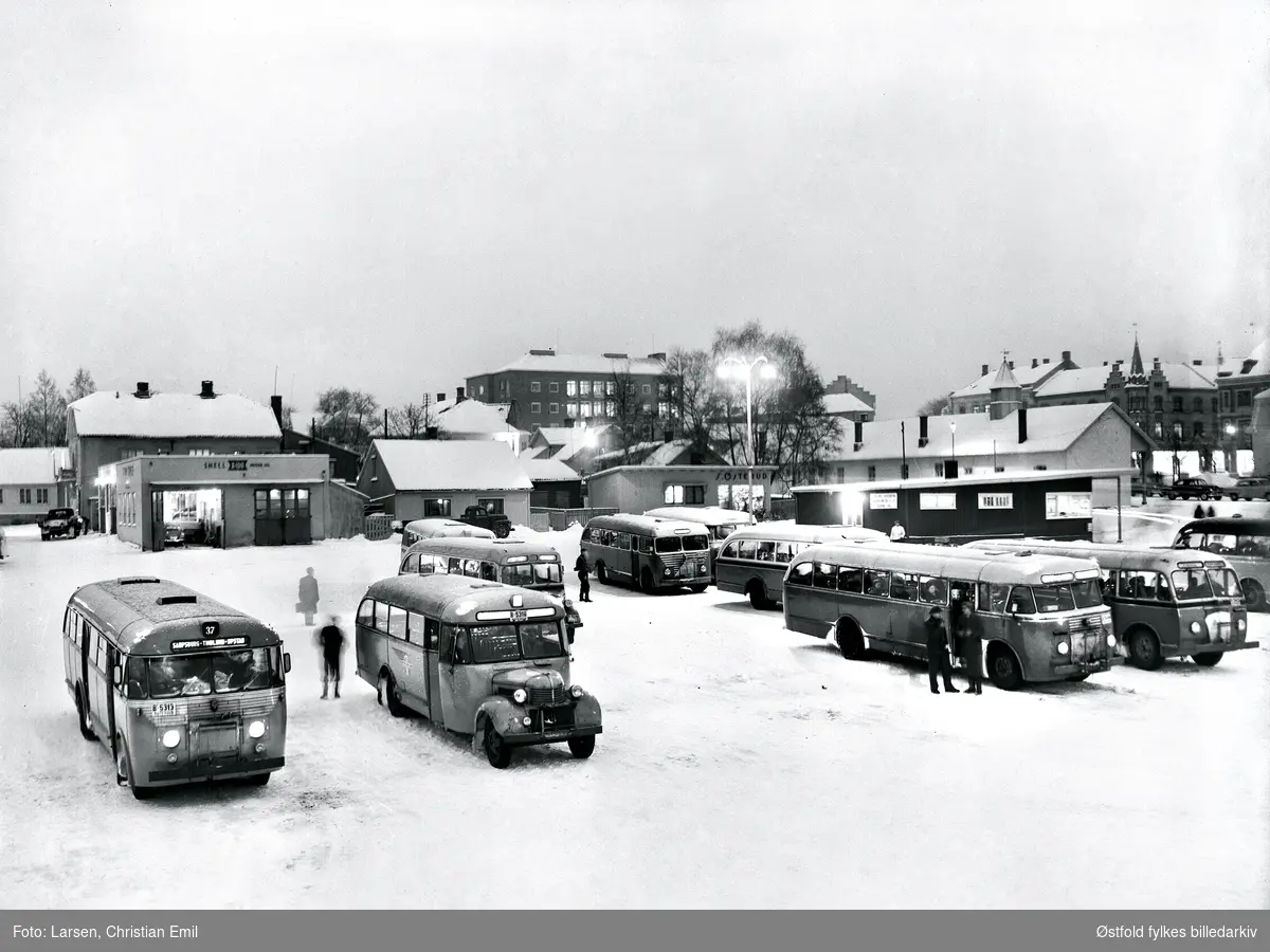 Busstorvet i Sarpsborg 1950-tallet, vinter og snø. Nedre torv, Sandtorvet. 
Forretning S. Østerud (kjøttbasaren) i bakgrunnen, og Shell bilverksted. Snutebusser og busser med flat  front, B-5316, B-5313.
Bussene skal være bygd hos Kristiansen og bussen helt til venstre er en en 1950-modell. Den mørke bygningen i bakgrunnen er Bussebua, kombinert venterom for passasjerer og sjåfører med en liten ekspedisjon (ekspedere gods), bussebua lå før på øvre torv. Bussebua skal være bygd 1940. Mannen mellom de to bakerste bussene er antakelig Buss-Ola, en byoriginal i Sarpsborg.