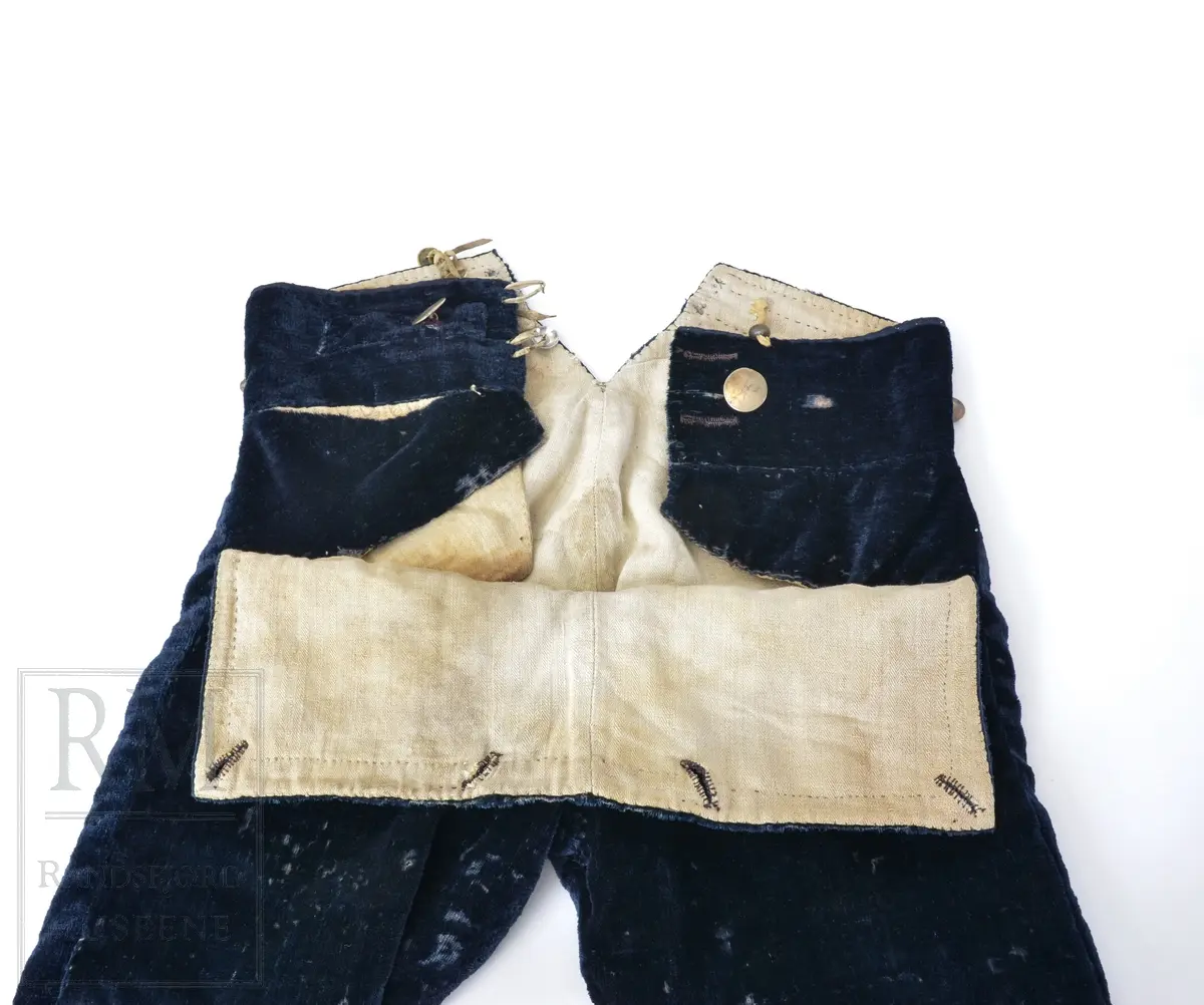 Mørk blå fløyelsbukse, knelang, med lem. Foret i lin. Fem flate messingknapper.