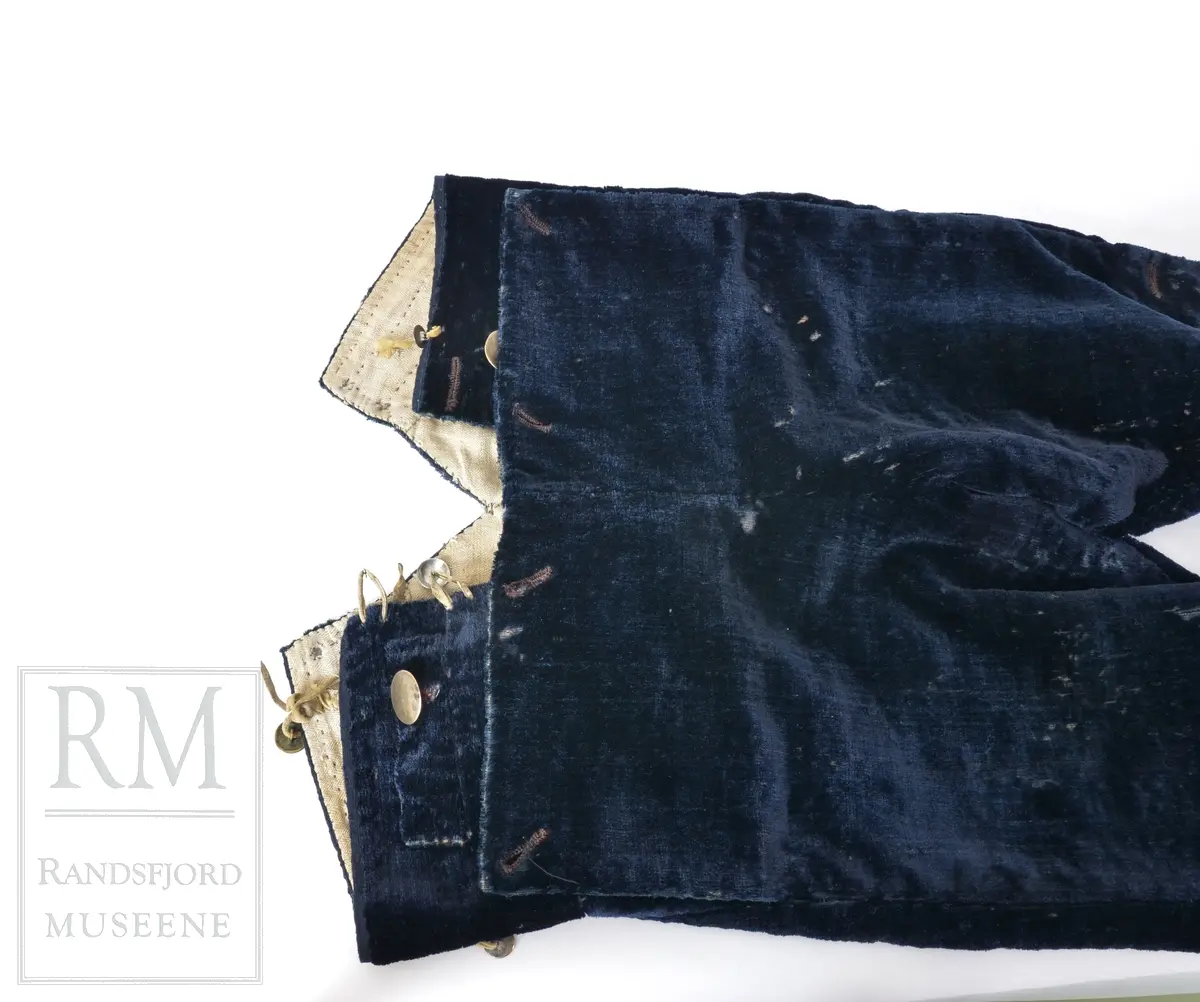 Mørk blå fløyelsbukse, knelang, med lem. Foret i lin. Fem flate messingknapper.