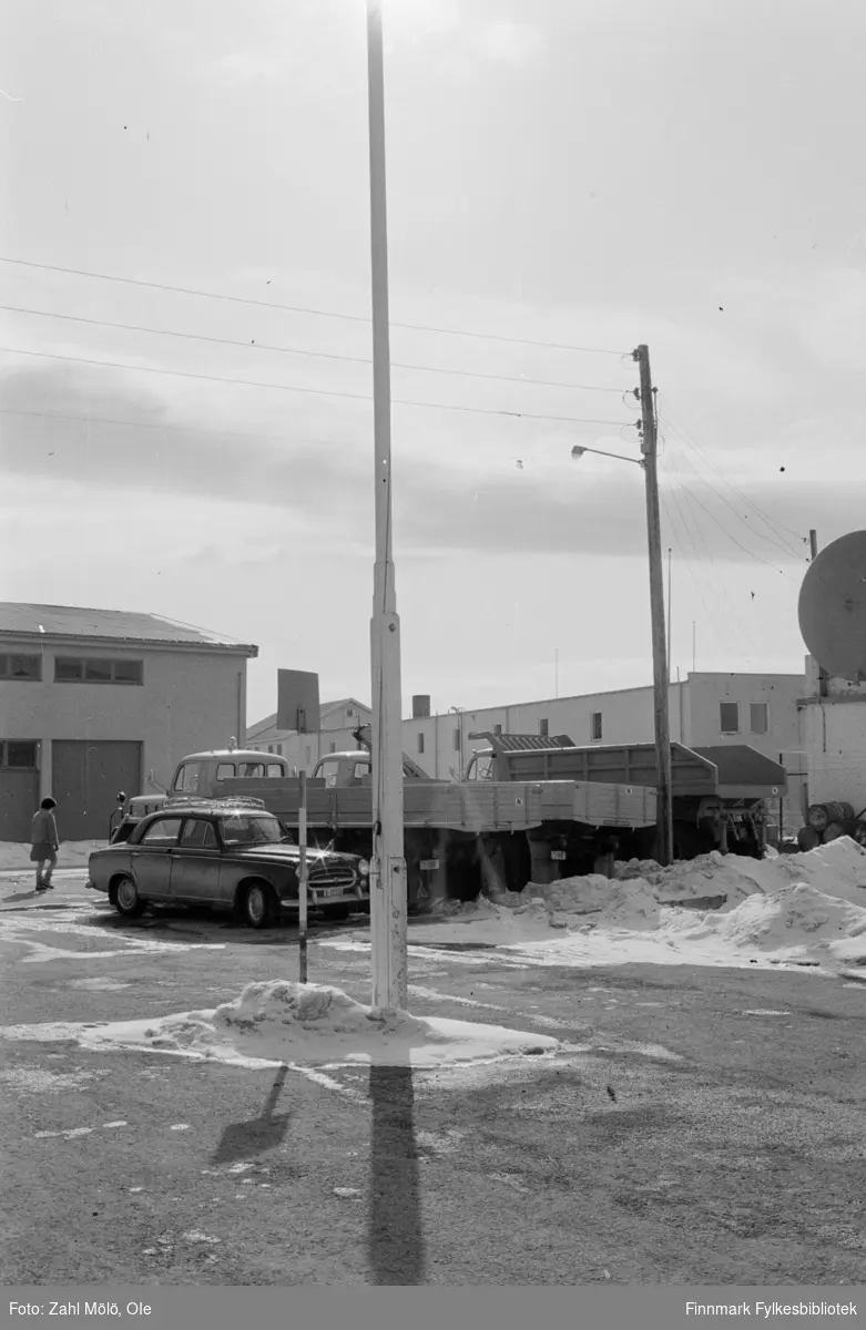 Fotoserie fra Vadsø, April 1968 fotografert av Vadsøfotografen Ole Zahl-Mölö. Vadsø by. En flaggstang ruver i front i bildet. Parkerte biler (nærmest er Peugeot 403) og lastebiler i bakgrunnen.