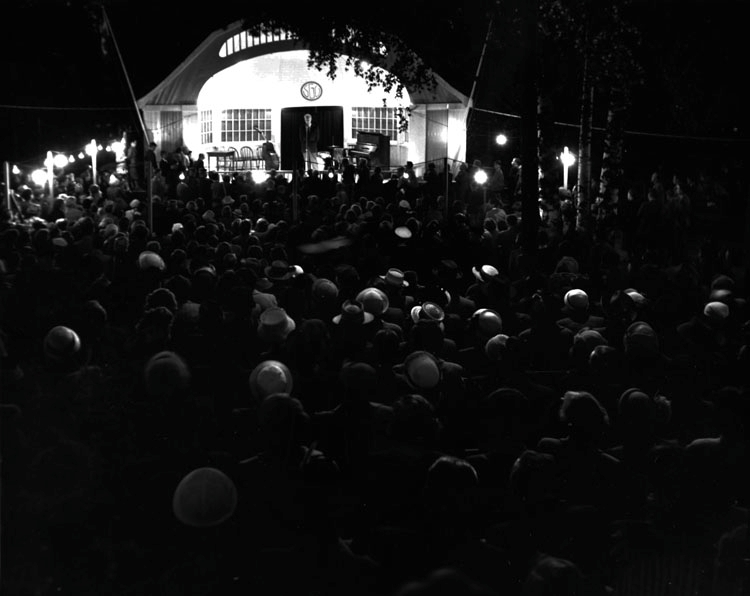 Kvällsfest på "Berget" i Söderhamn augusti 1953. En man står framför en mikrofon på en upplyst utescen med skylten SGU. Framför sitter en stor publik.