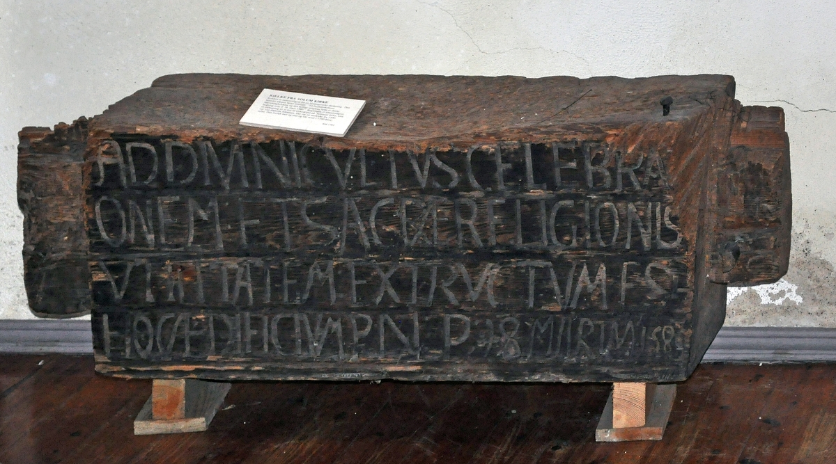 Fra protokollen:
Tømmerstok, rimeligvis fra en Bindingsværks døraapning af en ældre Kirke i Solum, nedrevet omtrent 1760, med følgende, i ophøiede Bokstaver, latinske indskrift: "ad divini cultus celebrationem et sacræ religionis utilitatam exstructum est hoc ædifiicium P. N. P. 18 marty 1580". Stokken er engang benyttet til huggeblok.

Tekst på skilt i gammel utstilling:
Bjelke fra Solum kirke
Stokken er sannsynligvis fra en bindingsverks døråpning. Den latinske teksten betyr oversatt: "Til Gudsdyrkelsens høytideligholdelse og den hellige religions nytte er denne bygning reist d. 18. mars 1580." Bygningen er sannsynligvis en påbygning av et kor til kirkeskipet på Soleimar kirke, som var viet døperen Johannes. Det var en stavkirke fra 1200-tallet. Den forfalt mer og mer og ble revet i 1760-årene.