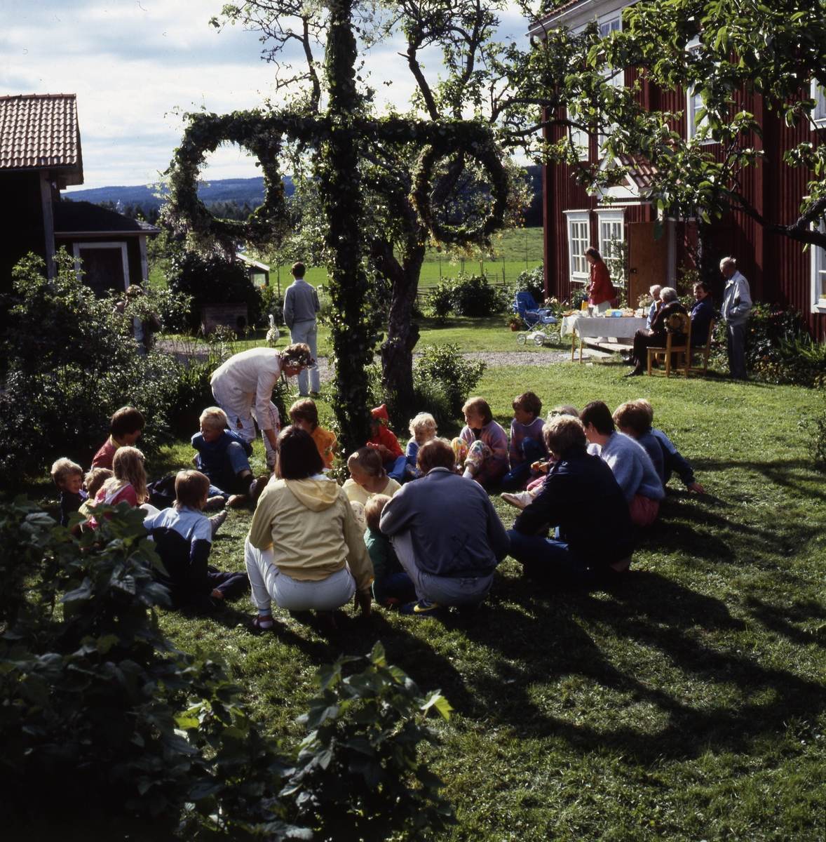 En grupp vuxna och barn sitter samlade på en gräsmatta runt en lövad midsommarstång. Vid entrén till huset sitter några vuxna vid ett bord, kanske dukat med fika.