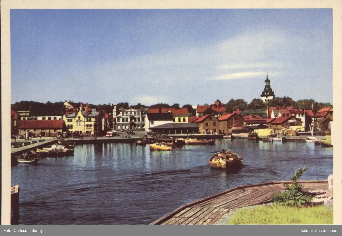 Fiskarhamnen i Västervik med småbåtar, framför staden med kyrka och byggnader. I förgrunden en brädklädd kajkant.