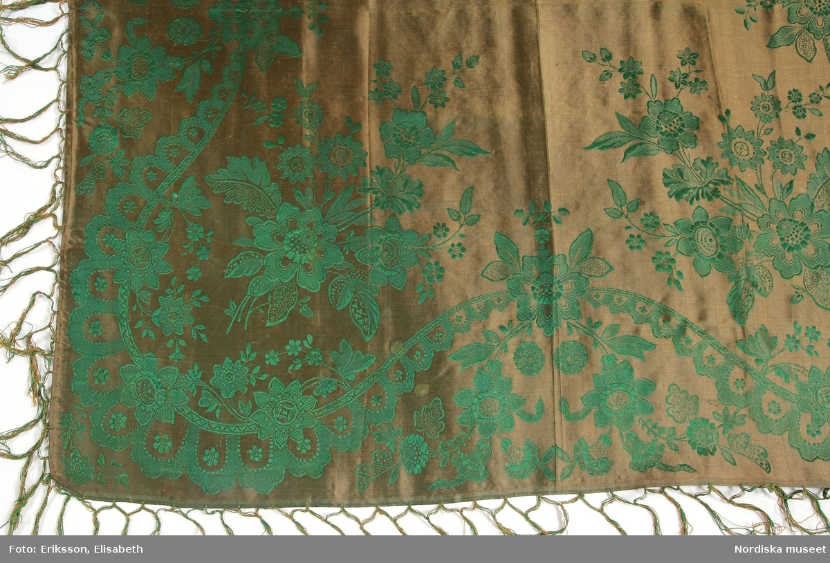 Kvadratiskt halskläde av sidensatin med jacquardvävt mönster, ljusbrun botten med grönt mönster, i varje hörn en stor blombukett, kantbård utan avgränsning med mjuk böjd slinga med uddkant och med utväxande blomkvistar. Runt om tvåknuten frans i klädets färger av tvinnat silke. I ena hörnet svart otydlig hallstämpel.
/Berit Eldvik 2011-11-