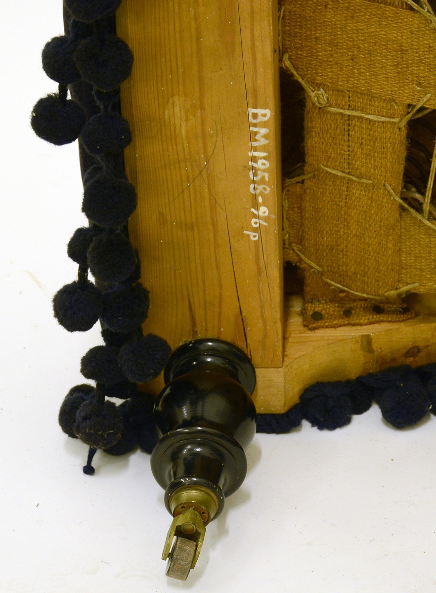 Ottoman med blått plysjtrekk og ponponger. Dreide ben i sortlakkert tre, hjul på føttene (et mangler). Tilhører møblement i tysk nyrenessansestil.