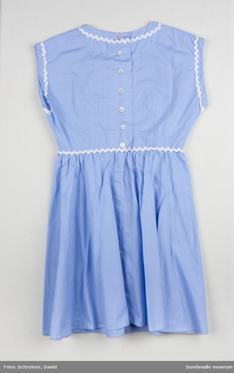 En ljusblå ärmlös klänning med utsmyckning runt ärmhål, halsrundning och midja med vitt dekorband.