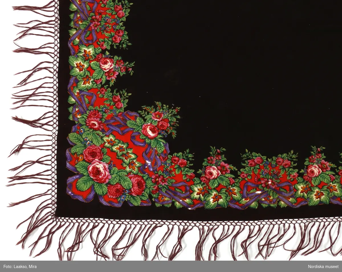Kvadratisk schal av mörkt brunviolett yllemuslin i kypert med tryckt bård runtom med bulliga rosor m.fl. mindre blommor och blad i rosa, blått, gröna nyanser samt ljusbrunt och vitt  med  stora anilinlila rosetter på klarröd botten. Färgerna mycket starka och klara och sannolikt anilinfärger av den sort som kom omkr. 1860. Invikta och tråcklade kanter på 3 sidor, en stadkant, treknuten frans av ullgarn i bottentygets färg. 
/Berit Eldvik 2012-01-31