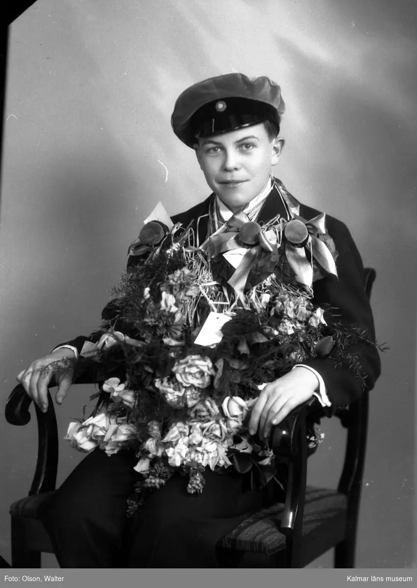 Ateljébild på en man som har tagit realexamen. Han har en mössa och blommor på sig, sittandes på en stol. Enligt Walter Olsons journal är bilden beställd av Ingvar Karlsson.