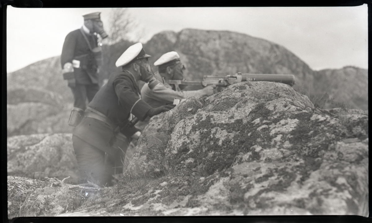 Tre militärer i gasmask bemannar kulspruta i skärgårdsterräng. Andra bilder i Bergers samling tyder på att detta fotografi tagits då manskap från pansarskeppet SVERIGE 1929 deltog i stridsövning på Stabbo i Stockholms södra skärgård, detta har dock inte kunnat bekräftas.