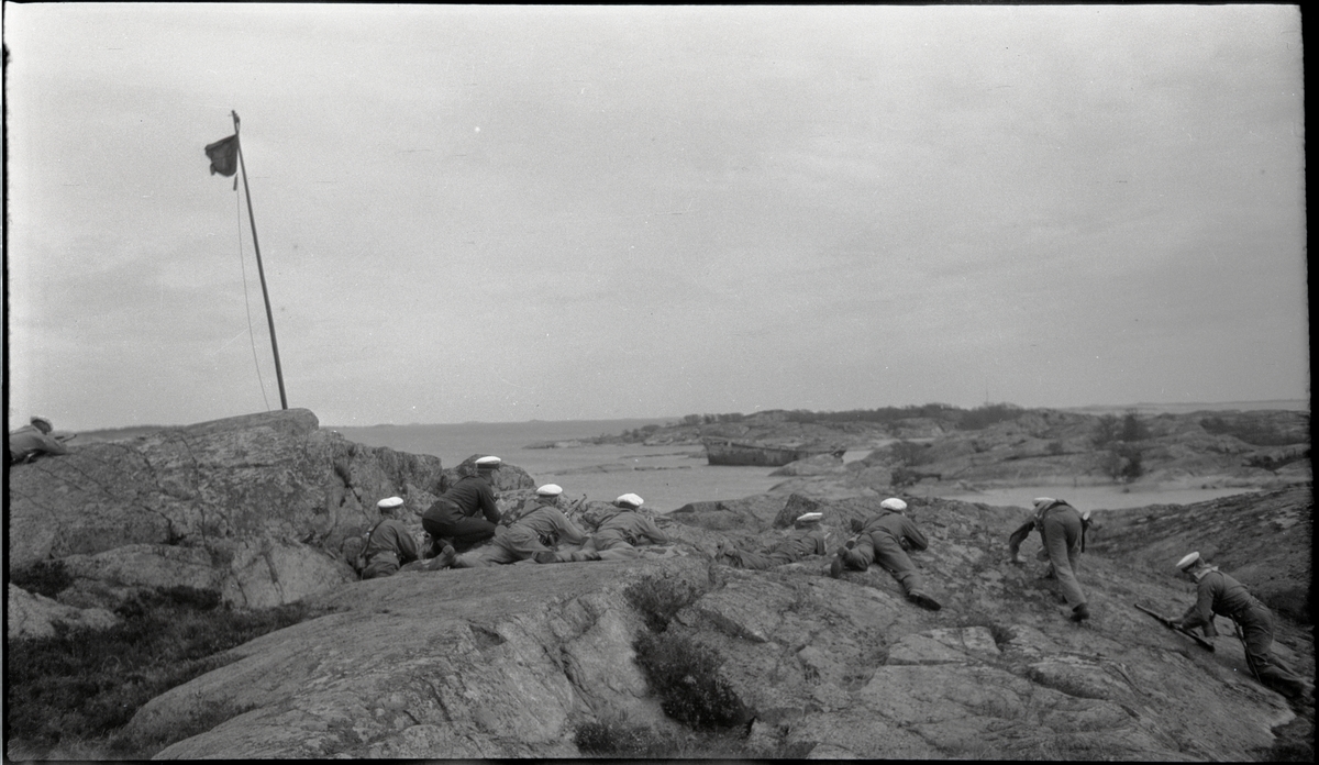Stridsövning på Stabbo i Stockholms södra skärgård 1929. Militärer med handeldvapen ligger på klipphällar och siktar mot fartygsvrak (skjutmål?) i bakgrunden.