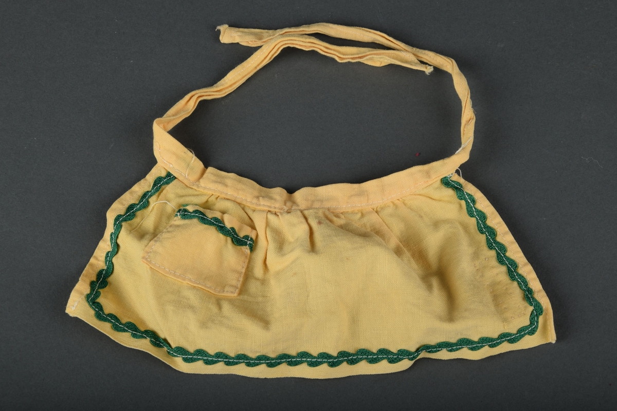 Forkle til dukke i gul tekstil. Liten lomme foran på forkleet og stropper til å knytte forkleet med. Påsydd grønt bånd langs kanten, og som detalje på lommen.