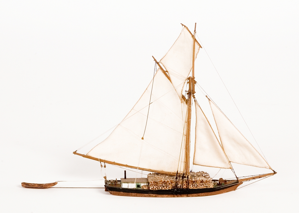 Fartygsmodell på vattenlinje. Roslagsslup, hel i block av trä med mast och segel av bomull. Svarta brädgångar, brunt skrov. Liten båt på släp.Föremålets form: Vattenlinjemodell