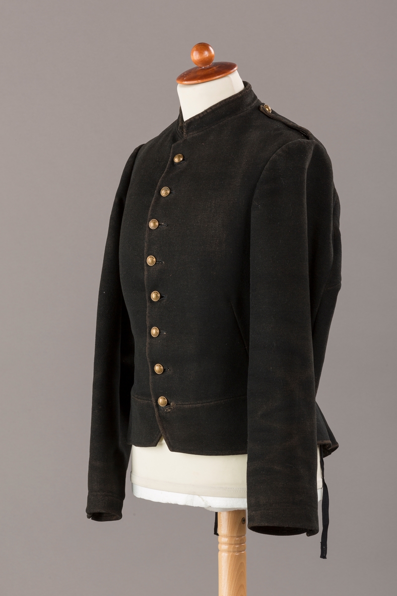 Form: Enkeltspent.

Foret med lin. Forsterket med silke på et lite stykke på ryggen. 12 messingknapper med mønster og tallet 1. Hempe i lin. Bendelbånd med knappehull.