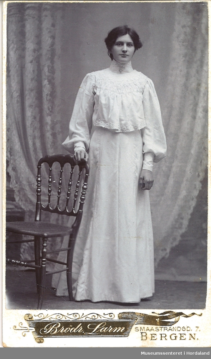 atelierfotografi av ung kvinne kledd i kvit brodert jakke og skjørt som stør seg på ein stol framfor draperte gardinar