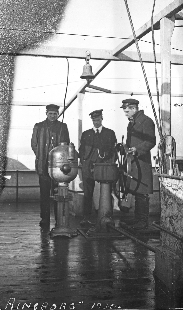 Mannskapsbilde. Tre av mannskapet på "Ringborg" samlet rundt skipsrattet på dekk. Hverdagsfoto.