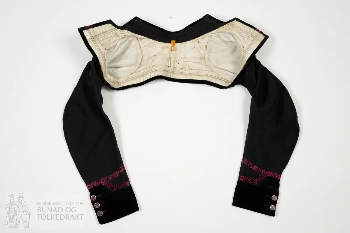 Opentrøye sauma av svart blank glatt klede, lerretsvove. Heilklipt rygg, bandprydd. Oppståande låg krage. Tosaumserme med bryningar. Open ermsliss med 3 stk knappar og knapphol til lukking. Bandpryding av blomstermønstra fløyelsband og svart bomullsfløyel til pryding av krage, framkantar, nedkantar og ermeslissar.  For av kvit litt glissen stiva bomullslerret. Maskinsaum og handsaum.