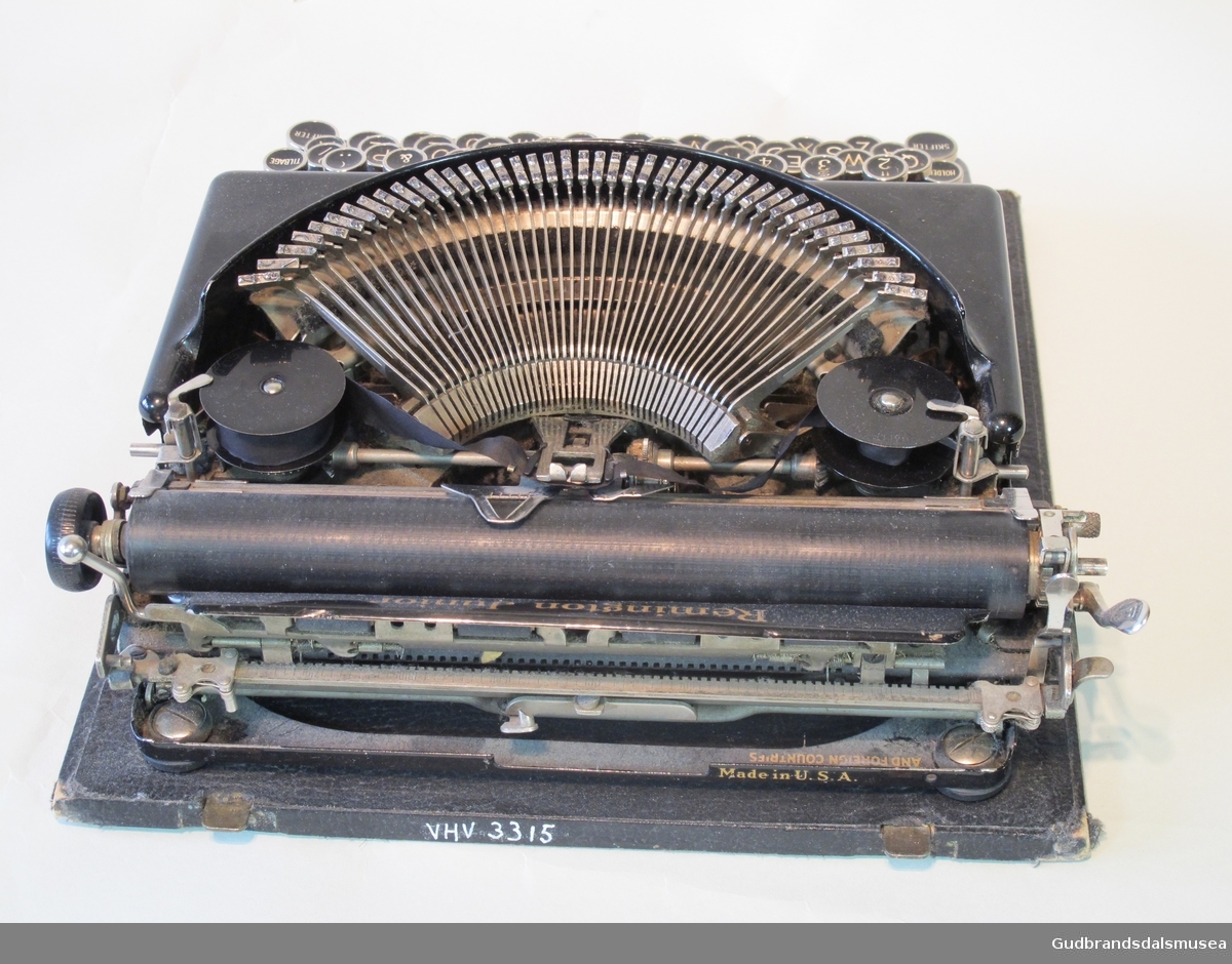 Manuell skrivemaskin med lokk. Har sort fargebånd. Norsk alfabet. Både maskin og lokk er noe slitt i kantene. Lokket har bærehåndtak.