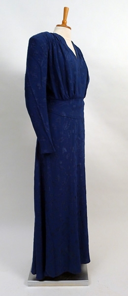 Enligt liggaren: Klänning dam av cloque, blå v-ringad, aftonklänning, draperad vid axlarna, blixtlås i sidan. Lång ärm axelvaddar. Midjebesparing, l 160 cm, bredd över axel 41 cm. 1940-tal