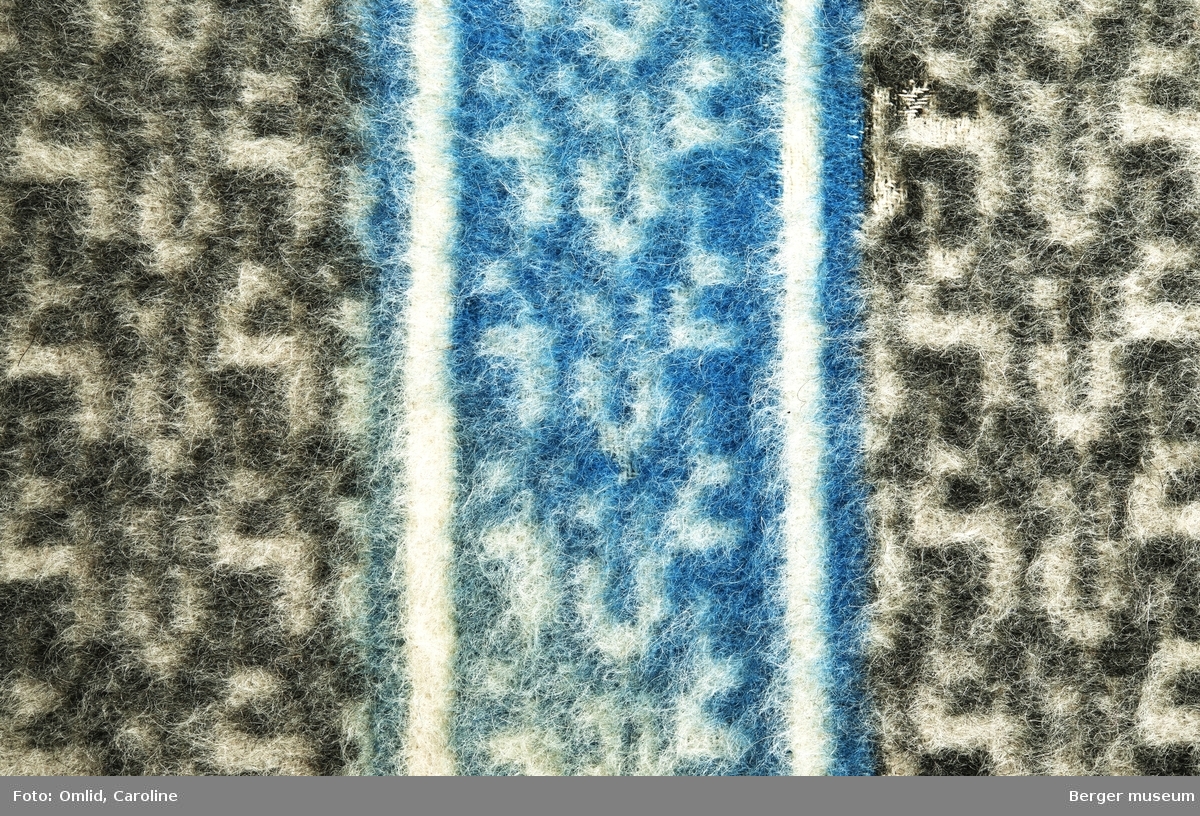 Teppe med geometrisk strukturmønster i mørk grått på lysere bakgrunn. Tverrgående blå bord med samme mønster.
