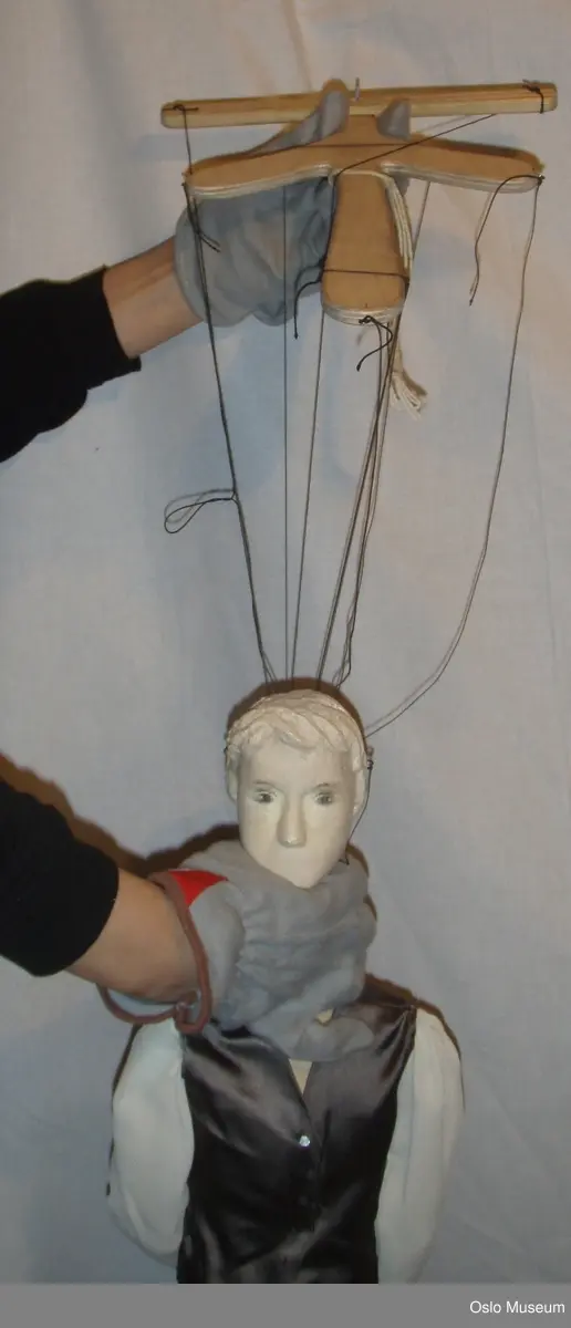 Marionett med karakter, ung mann.
Hodet er gips(?) og malt hvitt. Øyne er tegnet på. Har nese, men ingen munn. 
Halsen er i tre og er festet videre til overkroppen med et skinnsnøre som går ned i brystkassen og gjør hodet lett bevegelig. Overkroppen er i tre. Armene er bevegelig i skulder- og albueledd ved hjelp av noe tøy som er festet mellom treverket. Bena bevegelig i hofte- og kneledd.
Marionetten har på seg hvit bluse, der bare ermene er festet på ved skuldrene, en blank lilla vest med spiker til knapper og en lysere lilla, blank bukse som rekker til rett under knærne. Det er ni sorte tråder festet til kroppen: på knærne, hendene, korsryggen, skuldrene og på hver side av hodet. I den andre enden festes trådene til en  treholder som dukkeføreren bruker til å føre marionetten med.