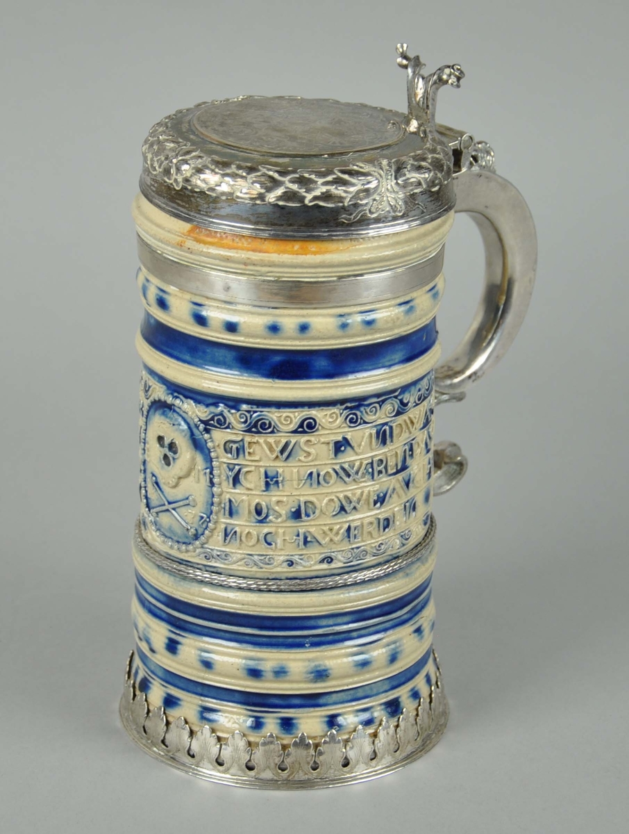 Stentøy ølkanne med sølvhank og sølvlokk. Hank og lokk er sekundære. På siden gjengitt et dødninghode med korslagte bein og en lengre innskrift på tysk. Sølvhank med dyremaske og sølvlokk med eikeløvskrans og gravert våpenskjold. Våpenskjoldet er familien Monrads våpen. Initiale FM og årstallet 1689 kan tyde på at det er Frederik Monrad, sogneprest i Øyer. Han var gift med Birgitte Cold. Initiale BCh kan være hennes, selv om navnet normalt ikke skrives med Ch.
Det tyder på at kannen opprinnelig har hatt hank av stentøy, som i 1689 er erstattet med sølvhank.