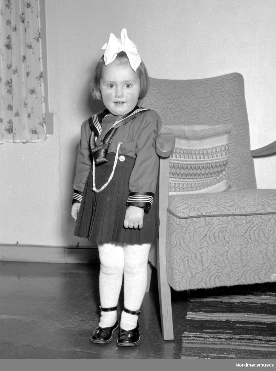 Foto av en liten pike stående ved siden av en stol. Bildet er registrert på Fridtjof Bjerkestrand. Fra Nils Williams fotoarkiv. Fra Nordmøre museums fotosamlinger.
