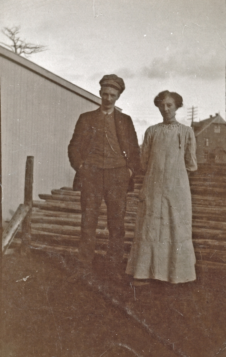 Mann og kvinne foran stabel med tømmer, ant. butikkmedarbeidere hos Meyer. Bygning bak.