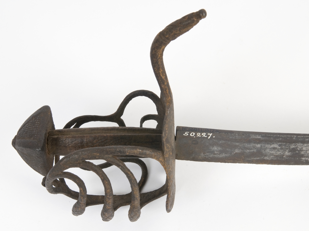 Pyramideformet  knapp med punslet strekdekor. Tregrep. Runde parerstenger med filt strekdekor og svakt nedbøyd bakre parestang. Fremre parerstang er brukket. Kurvfeste med runde bøyler med utsmidde, skråfilte midtpartier. Nyreformet parerplate med tombøyle på innsiden. 
2 sidebøyler på innsiden.
Enegget, kileformet krum klinge med  ryggstilt blodrand. Gjeddenebb.  
Klingens innside merket med ett smedstempel.