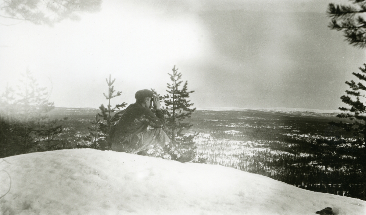 Mann med kikkert sittende på Buskneshøgda, Galåsen, og speider utover mot grensefjellranda i horisonten.