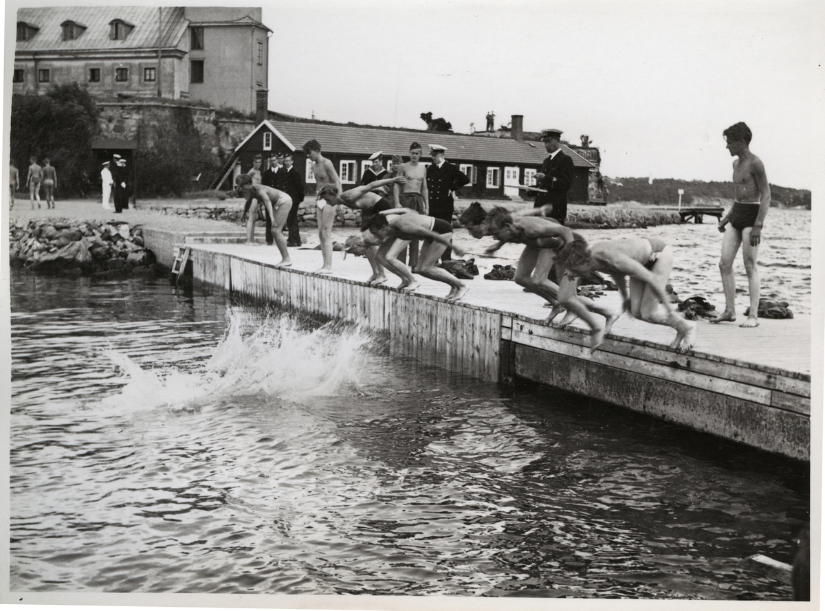 Bilden visar en grupp värnpliktiga under simutbildning som håller på att hoppa i havet från en brygga. Flera officerare övervaka och anteckna skendet. I bakgrunden syns Kungshall fästning på Stumholmen i Karlskrona.