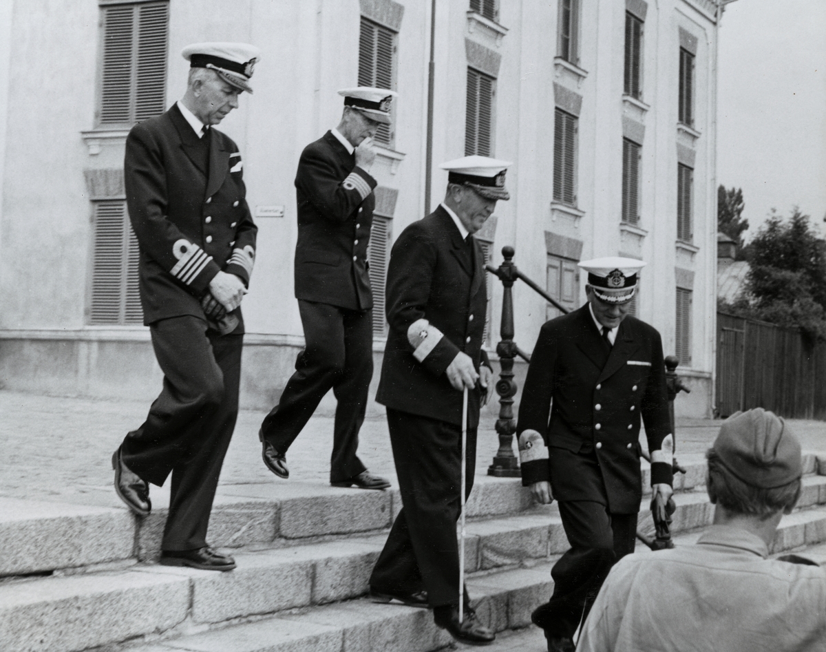 Konteramiral Samuelson (andra från höger) och kommendörkapten Lind af Hageby (andra från vänster) går ned för en trappa vid Kungsbron tillsammans med ytterligare en konteramiral och en kommendör. Samtliga är klädda i uniform. I bakgrunden syns Hollströmska magasinet.