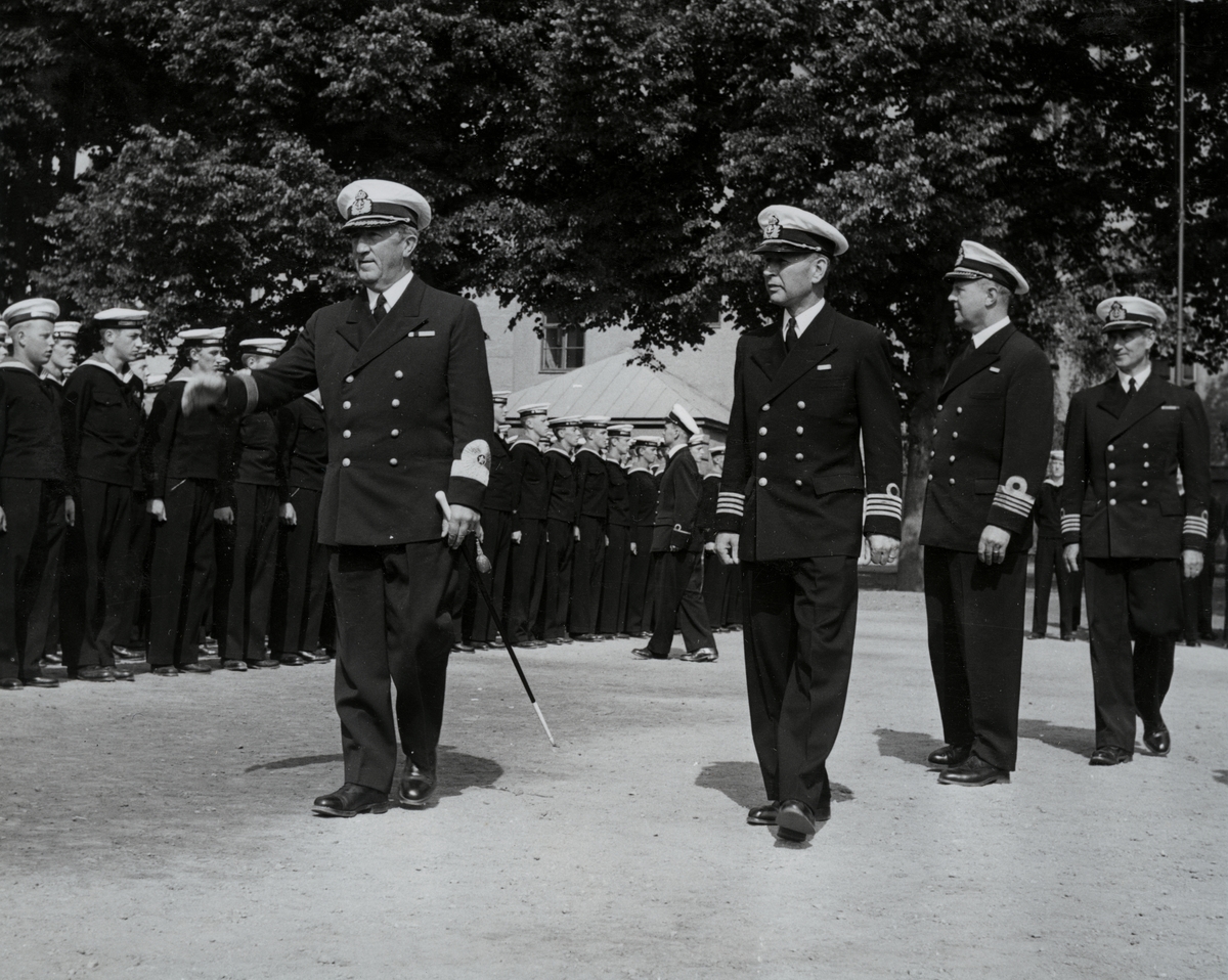 Konteramiral Samuelson (till vänster) och kommendörkapten Lind af Hageby (till höger) går, tillsammans med kommendör Bong och en kommendörkapten, förbi uppställda flottister. Samtliga är klädda i uniform.