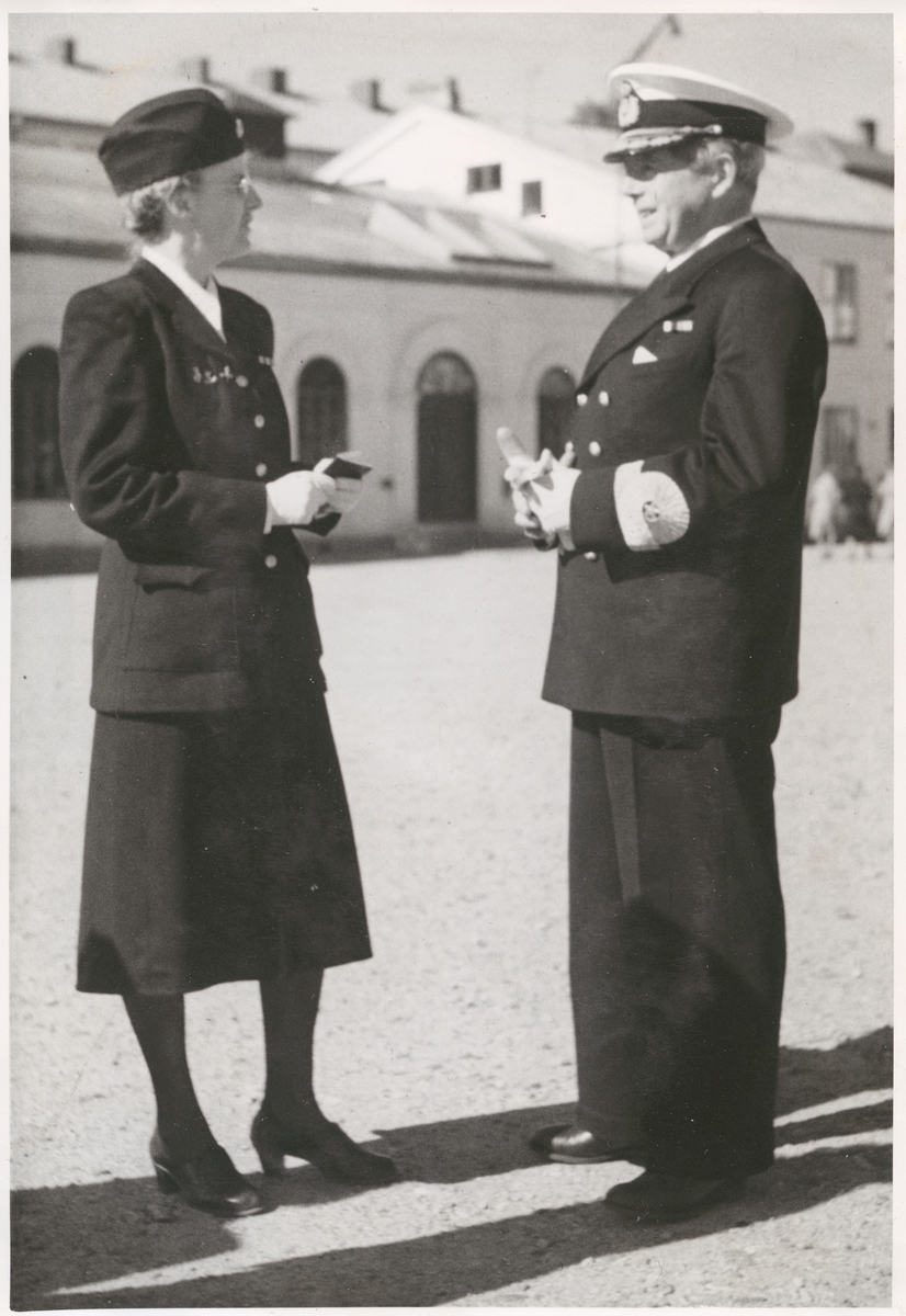 Konteramiral Gösta Ehrensvärd står på bataljon Sparres kaserngård och samtalar med en lotta. Båda är klädda i uniform och mössa. I bakgrunden syns exercishuset tillhörande bataljon Sparre, med putsad fasad och rundbågiga fönsteröppningar.