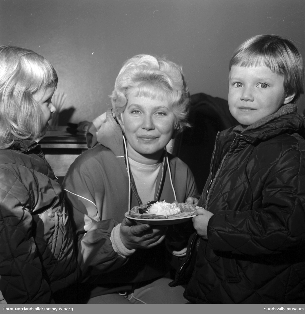 Inför våffeldagen poserar en kvinna och två barn med en våffla med sylt och grädde på ett fat.