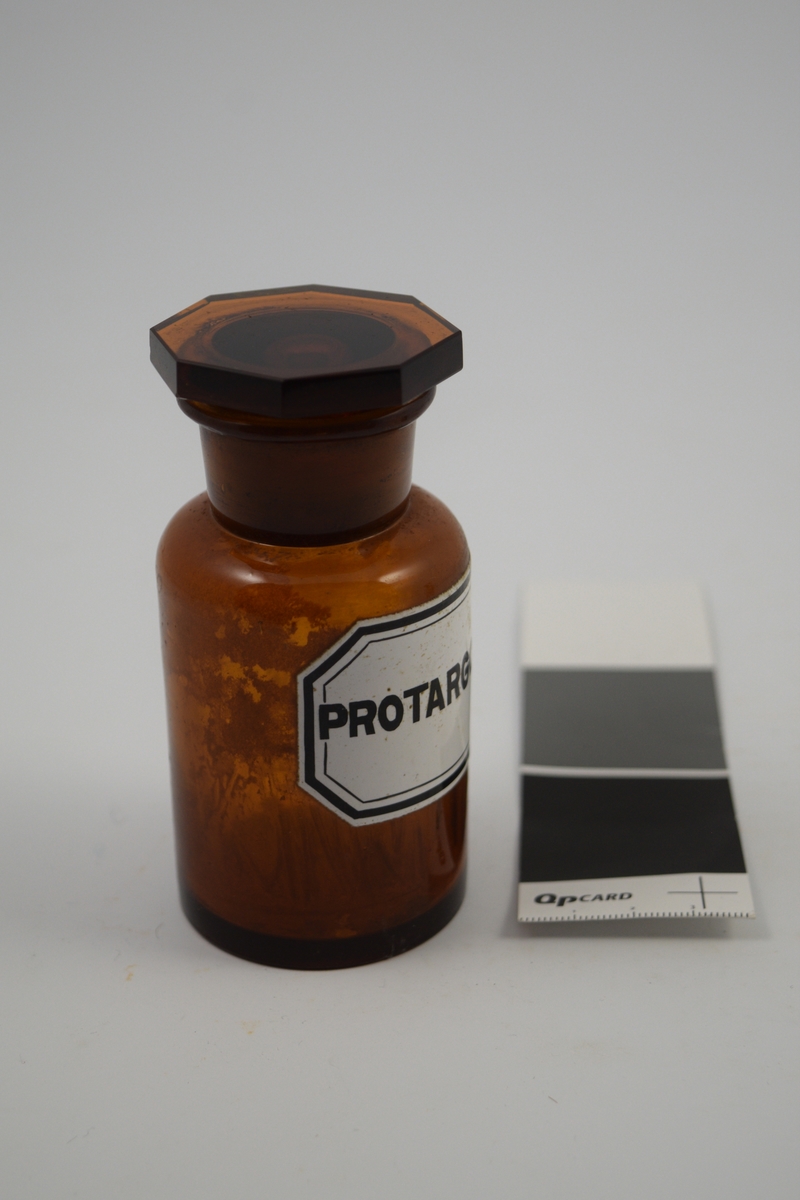 Brun glasskrukke med brun propp. Hvit etikett med sort skrift. Brukt til oppbevaring av Protargolum (sølvproteinat), som tidligere ble brukt som nesedråper.