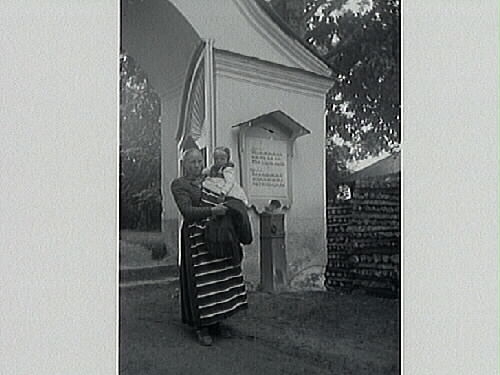 Kvinna iklädd sockendräkt med ett barn på armen står framför stigport till kyrkogård. Vid stigporten står en fattigbössa med en tavla över där texten ber om gåvor till de fattiga. Till höger i bild syns en vedtrave.