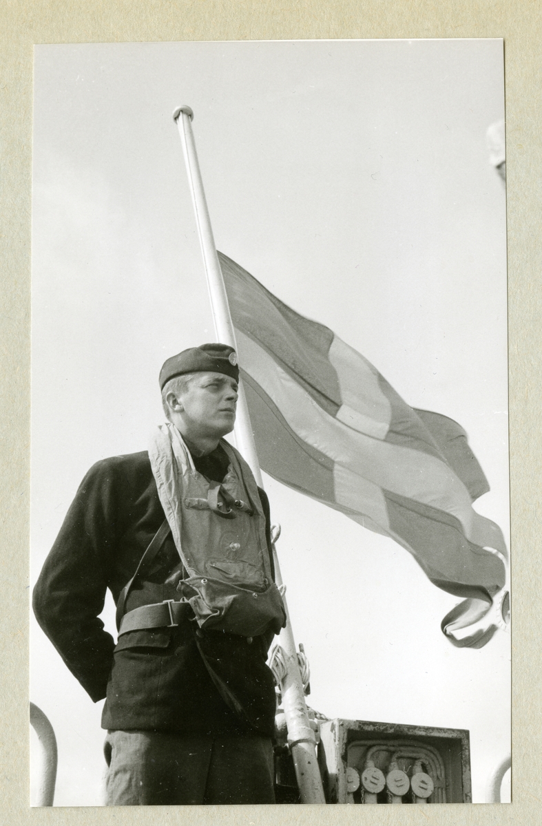 Bilden föreställer en man i uniform som står vid en svensk flagga som vajar i vinden på halv stång. Bilden är tagen i samband med en minnesgudstjänst under minfartyget Älvsnabbens långresa 1966-1967.