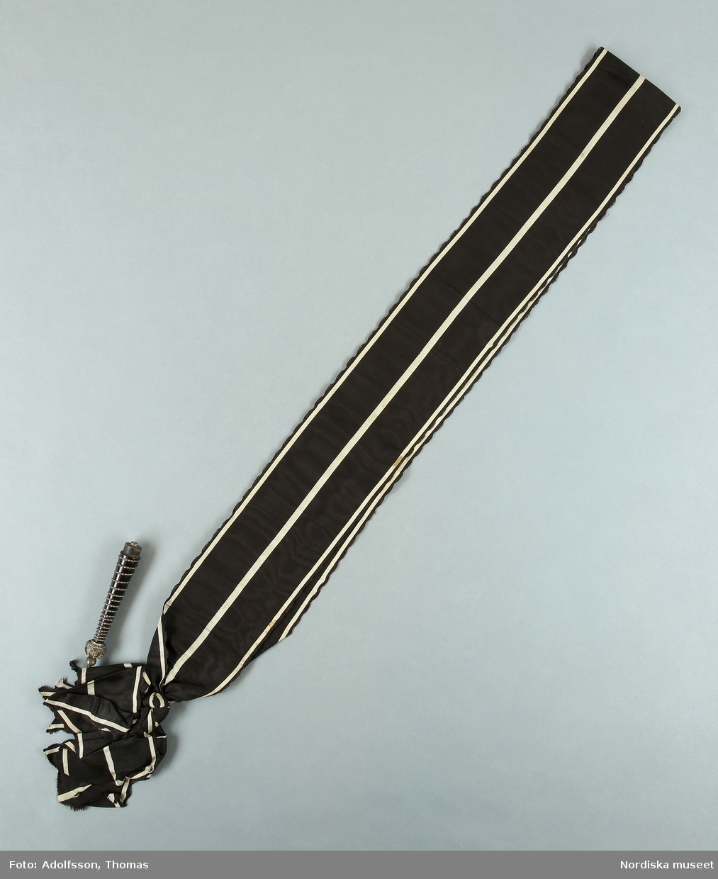 Huvudliggaren 1896:
"d. L. 0,13 Battong af svart trä, lindad med hvit metalltråd samt med kunglig krona af hvit metall.
I svart sidenmoaréband med hvita ränder. Bars öfver axeln."