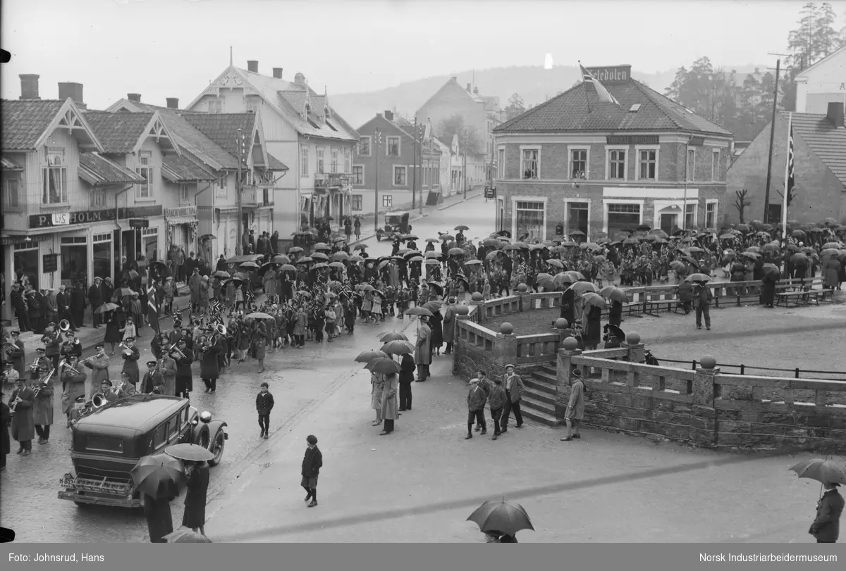 17. mai feiring 1931. Folketog med flagg og korps i gaten ved Notodden torg. Tilskuere på fortau med paraplyer. Biler og mennesker gående i gaten. Teledølen lokale i bakgrunnen. Butikkfasader langs Storgata i Notodden sentrum.