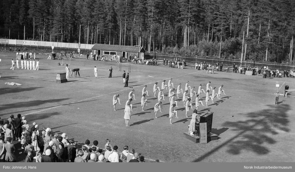 Turnstevne 1933. Turndamer har oppvisning, to kvinner ved piano stående på gresset. Menn som utøver turn på apparater ved siden. Tilskuere rundt stadion.