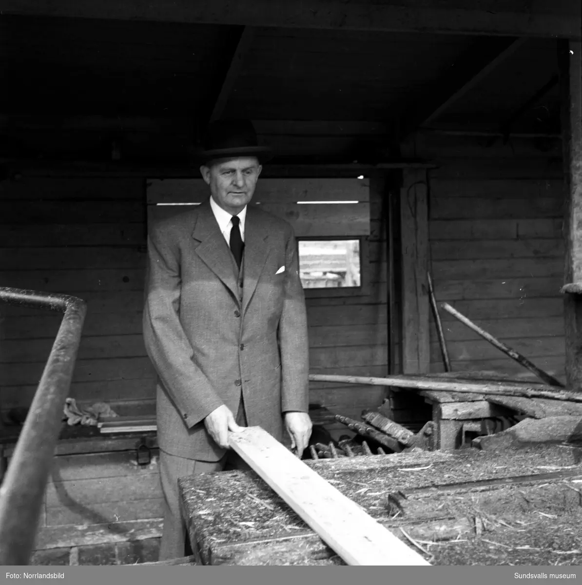 En man i hatt, kostym och slips pratar med en arbetare i såghuset på Skönviks sågverk.