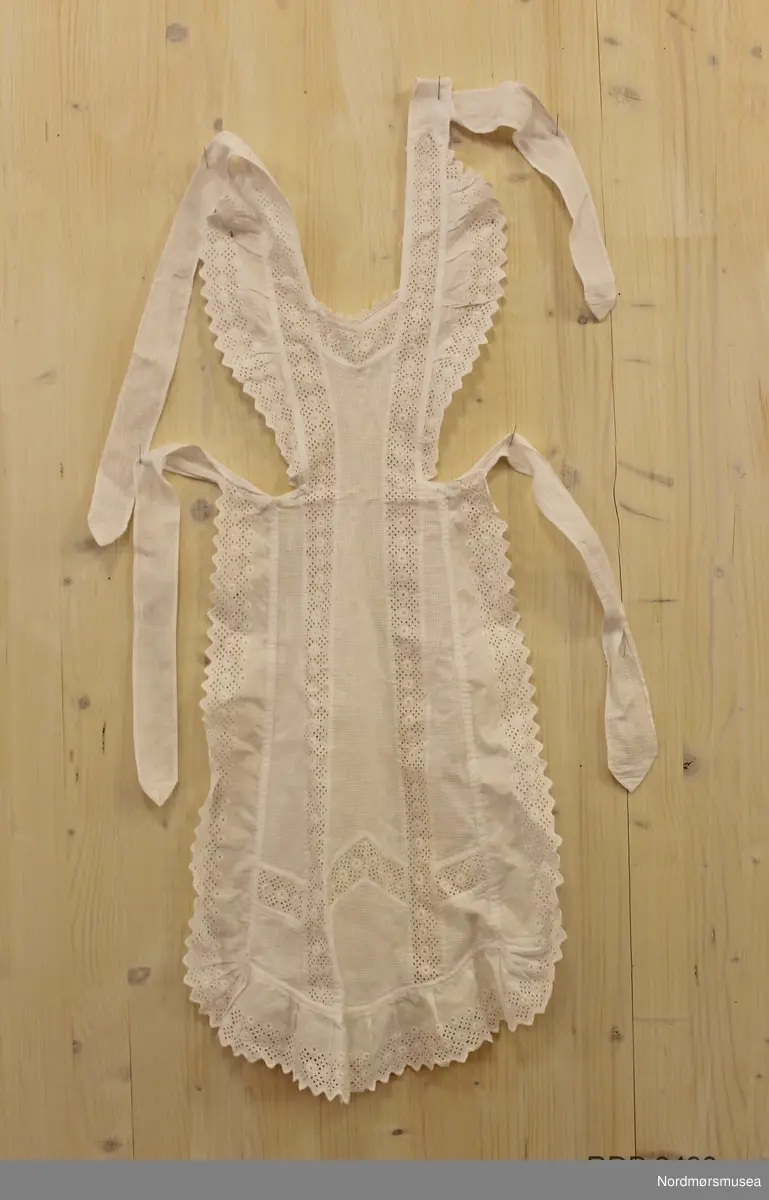 Kvitt serveringsforkle i bomull med innfeldte blonder og blondekant rundt. Smal midje og smekke. Knytteband rundt nakken og midja. Maskinsydd.