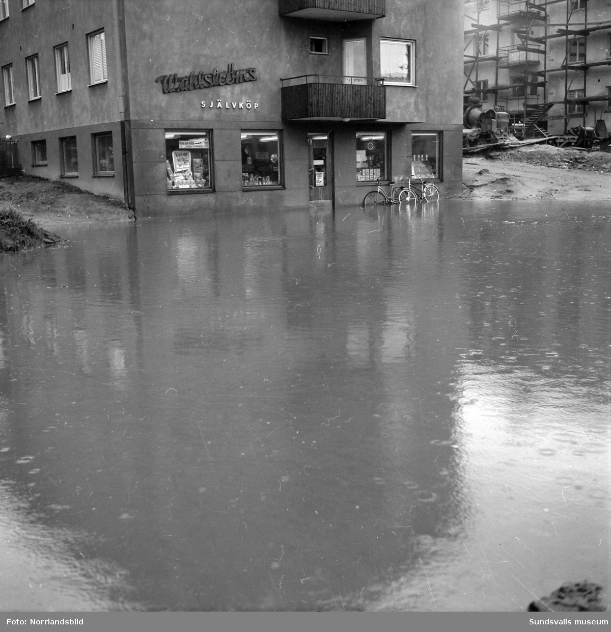 Översvämningar i Sundsvall, de flesta bilderna från Björneborgsgatan 11-24 samt Grevenbäcken som svämmat över. Wahlströms självköp och Städernas Vakt AB har råkat illa ut och källarvåningarna i flerfamiljshusen är drabbade. Vid Aluminiumkompaniet har en översvämmad bäck skurit av landsvägen och bildat ett vattenfall. På en av bilderna ses fyra män leta efter en avloppstrumma i Grevebäcken stånde dels i en båt och dels på en omkullvräkt arbetarkur. Det är förman Jonsson, E. Forsberg, G. Persson och A. Söderman.