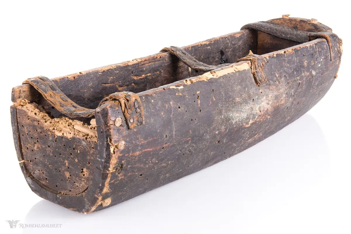 Uthulet trestokk som er høvlet flat i bunnen og har påsatte endestykker som er festet med trenagler. Tre lærremmer er spikret fast på tvers av åpningen. Rester av bek i bunnen.