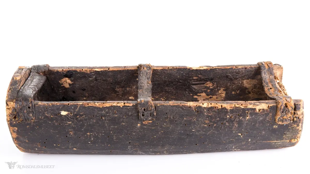 Uthulet trestokk som er høvlet flat i bunnen og har påsatte endestykker som er festet med trenagler. Tre lærremmer er spikret fast på tvers av åpningen. Rester av bek i bunnen.