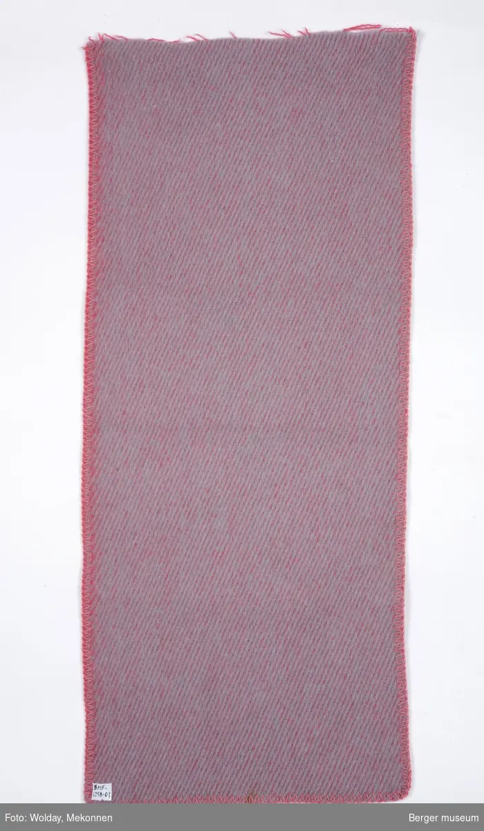 Et avlangt stykke pleddprøve, som er klippet av i ene enden, de andre sidene er sydd rundt. Teppet er to-farget, emd en farge på hver side.
